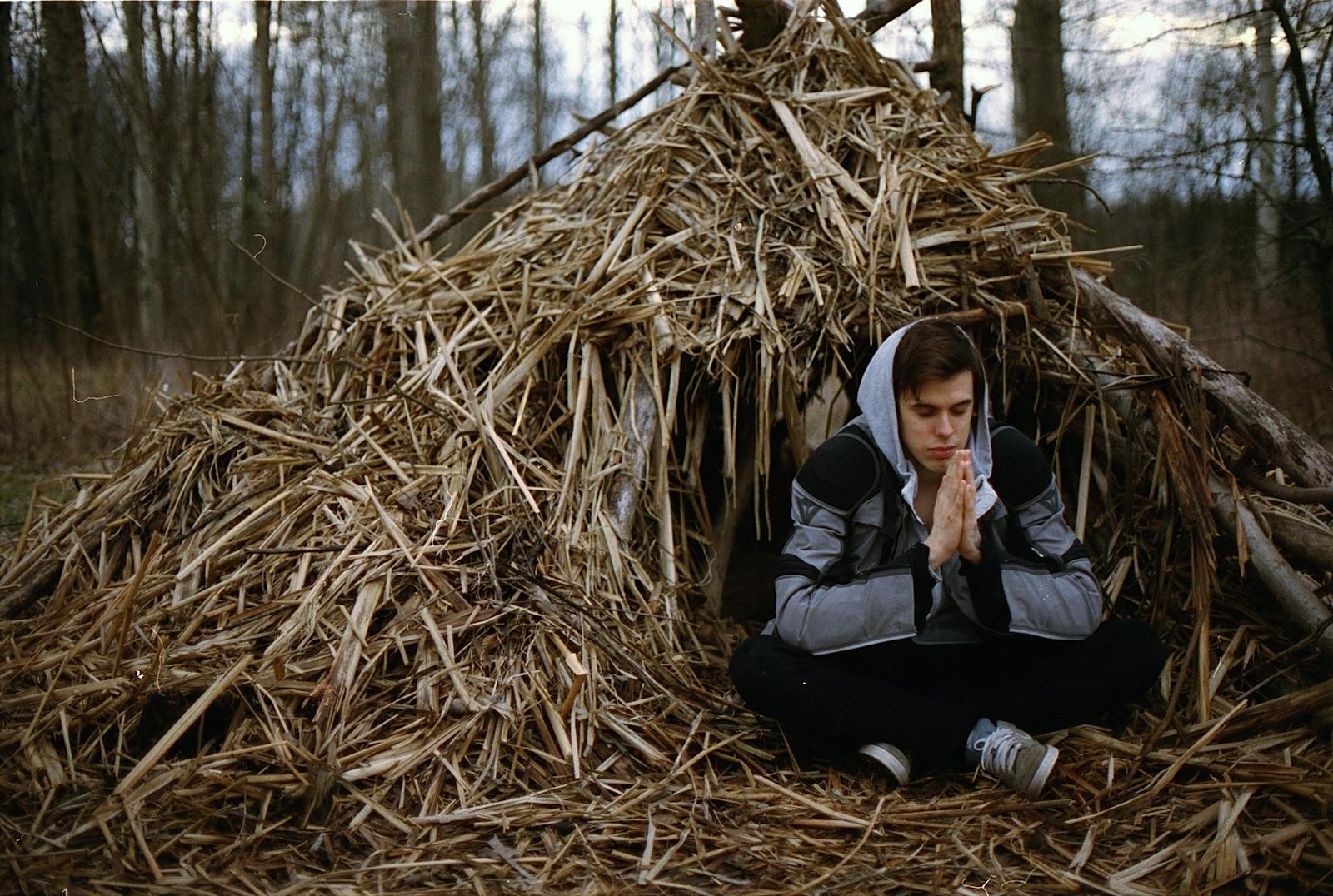 oración de la mañana, Adult male meditating outdoors by a rustic hay hut in a serene forest setting.