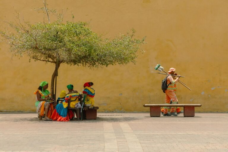 Oración por la paz de Colombia, Women in vibrant traditional attire rest under a tree while a worker passes by.