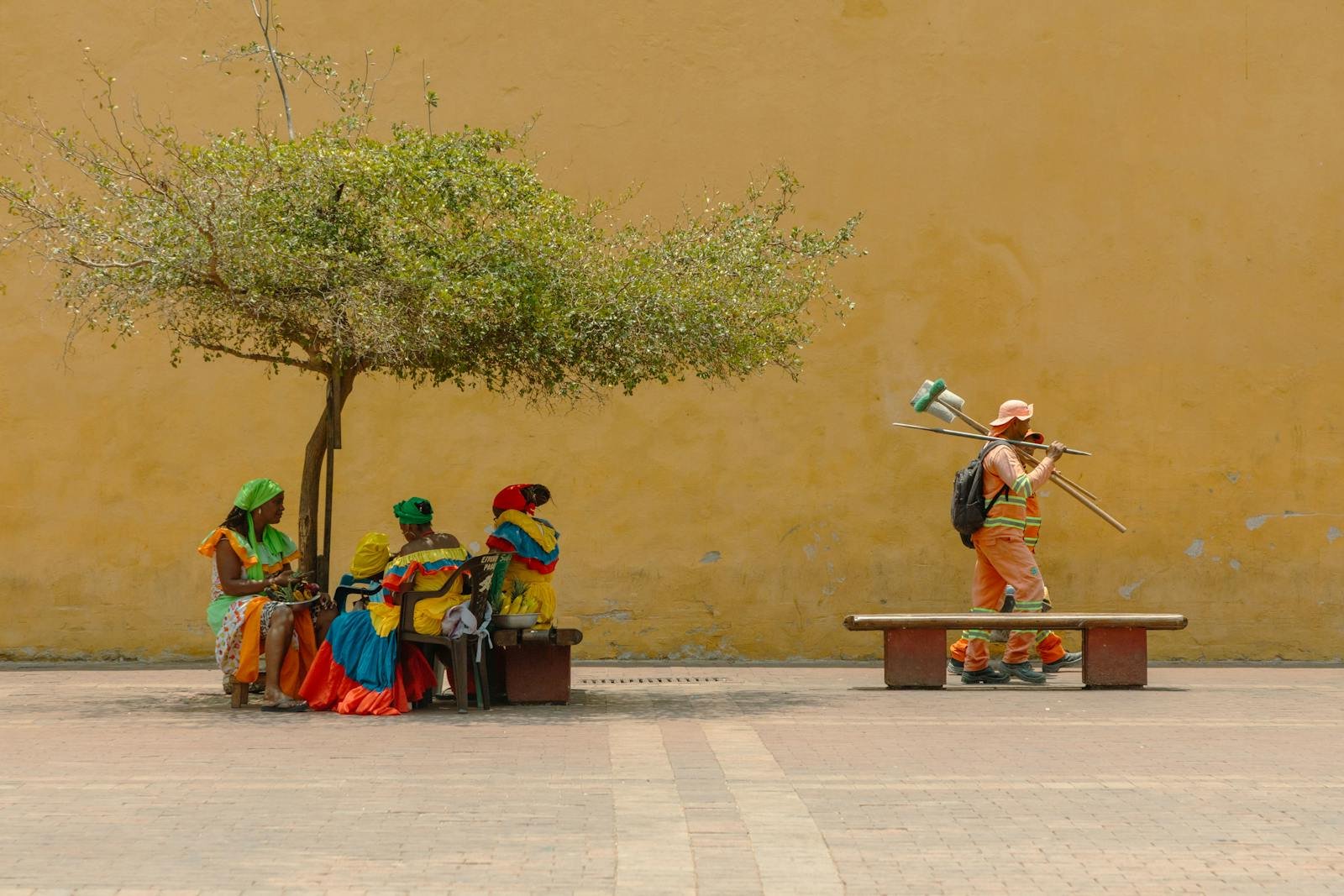 Oración por la paz de Colombia, Women in vibrant traditional attire rest under a tree while a worker passes by.