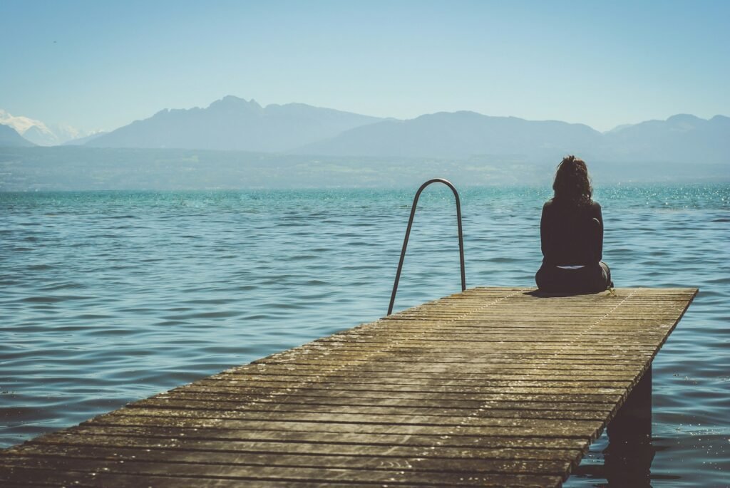 para qué estoy en este mundo, a woman sits on the end of a dock during daytime staring across a lake